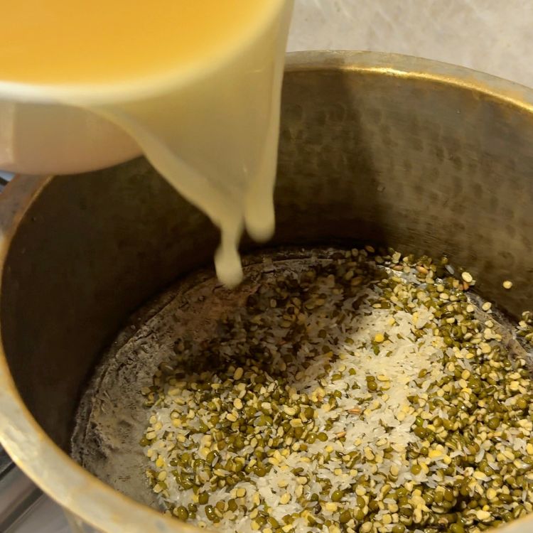 Chicken broth being poured into a pot with rice and split green moong dal on a stovetop.