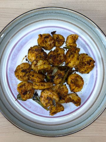Crispy golden Indian spiced shrimp fry served on a white plate with fresh curry leaves garnish on a wooden table