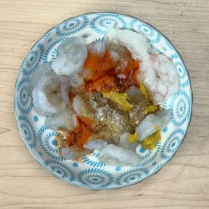 Raw peeled and deveined shrimp in a bowl next to small bowls of red chili powder, cumin, garam masala, coriander powder, and ginger garlic paste