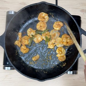 Hands using a wooden spatula to stir spiced shrimp as they cook and turn pink in a frying pan