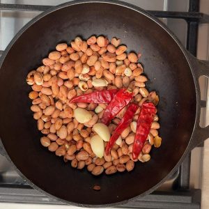 Red chilies and garlic cloves being added to golden roasted peanuts in pan