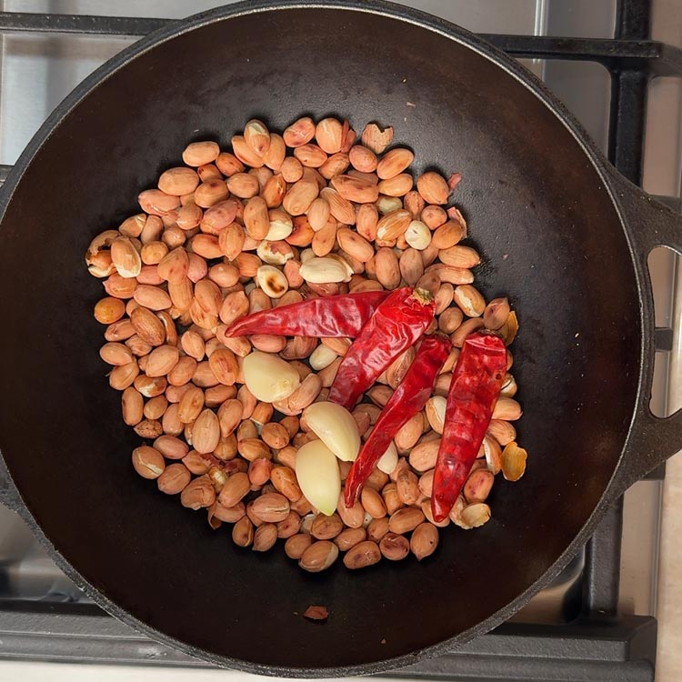 Red chilies and garlic cloves being added to golden roasted peanuts in pan