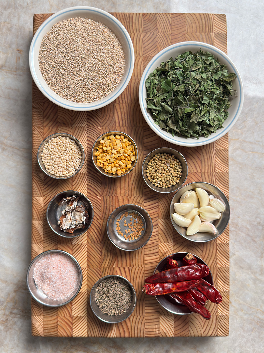 Raw ingredients for podi recipe arranged on wooden surface including white sesame seeds, curry leaves, and spices