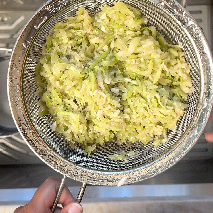 Cooked vegetable being pressed with spoon in metal strainer to remove excess water