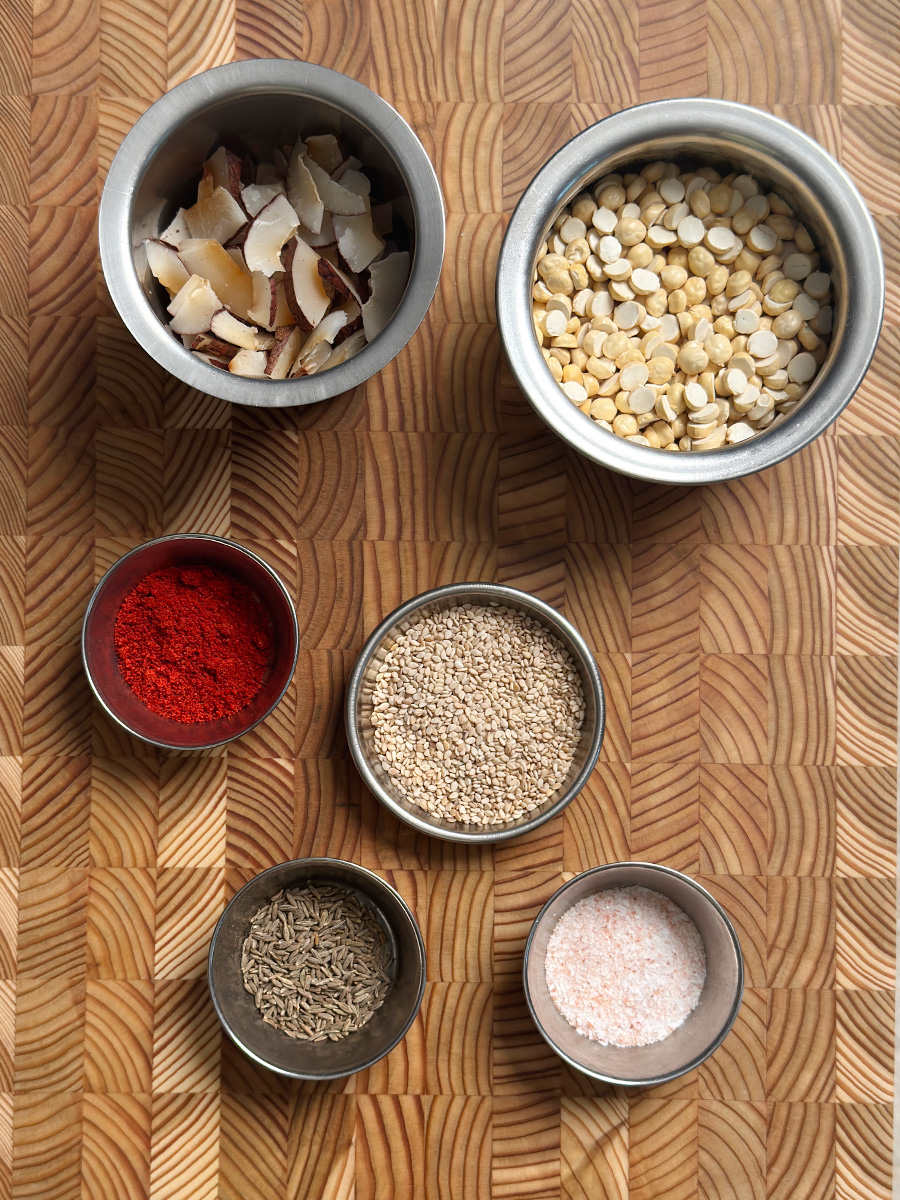 Roasted chana dal, dried coconut slices, cumin seeds, red chili powder, and sesame seeds arranged in small bowls