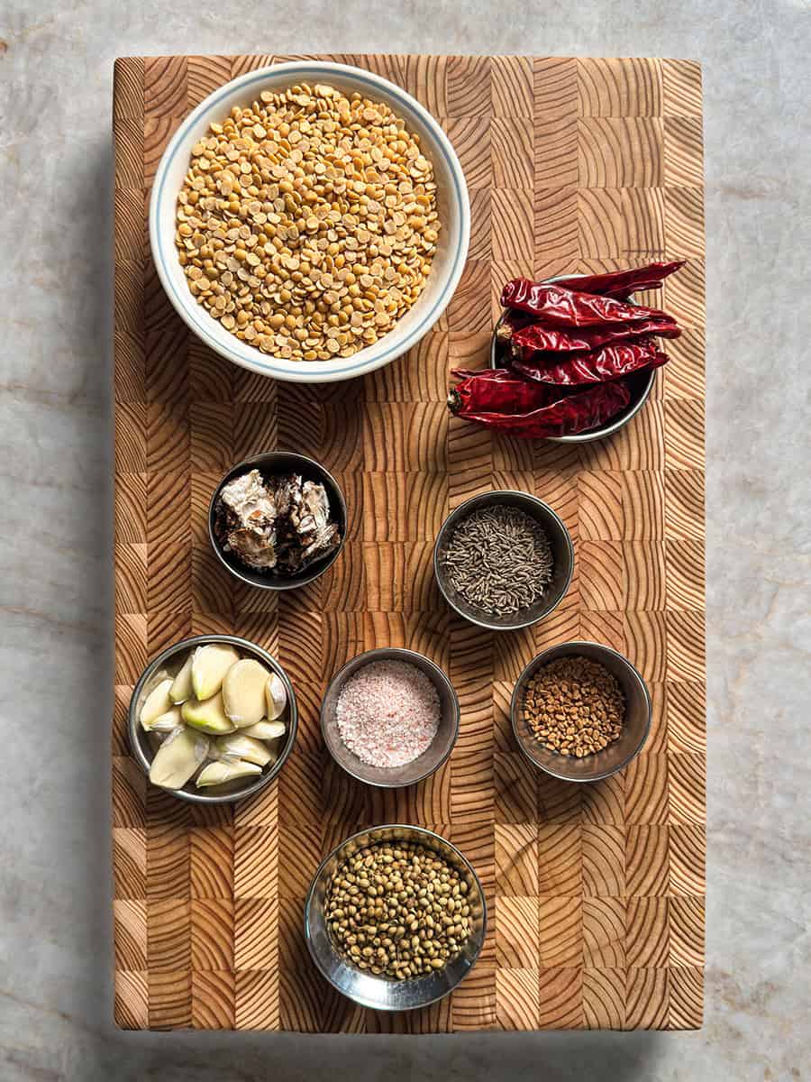 Toor dal, tamarind, cumin seeds, coriander seeds, fenugreek seeds, dried red chilies, garlic, and salt arranged on wooden table