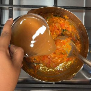 Dark brown tamarind water being poured into the spiced tomato gravy creating a tangy base