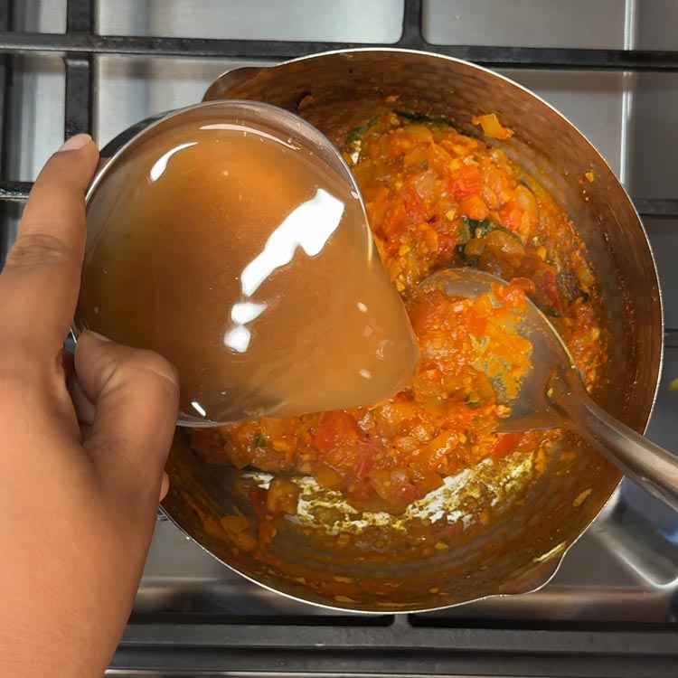 Dark brown tamarind water being poured into the spiced tomato gravy creating a tangy base