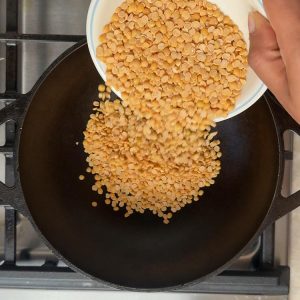 Golden split pigeon peas being poured from measuring cup into black cast iron skillet