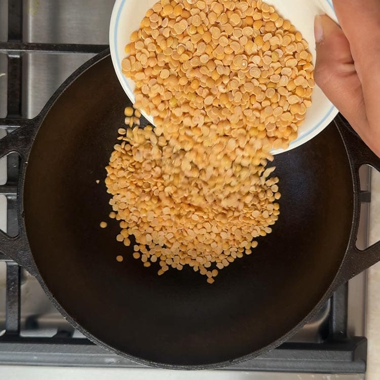 Golden split pigeon peas being poured from measuring cup into black cast iron skillet