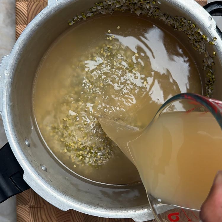 Pouring golden chicken broth from glass measuring cup into pressure cooker with rice and dal