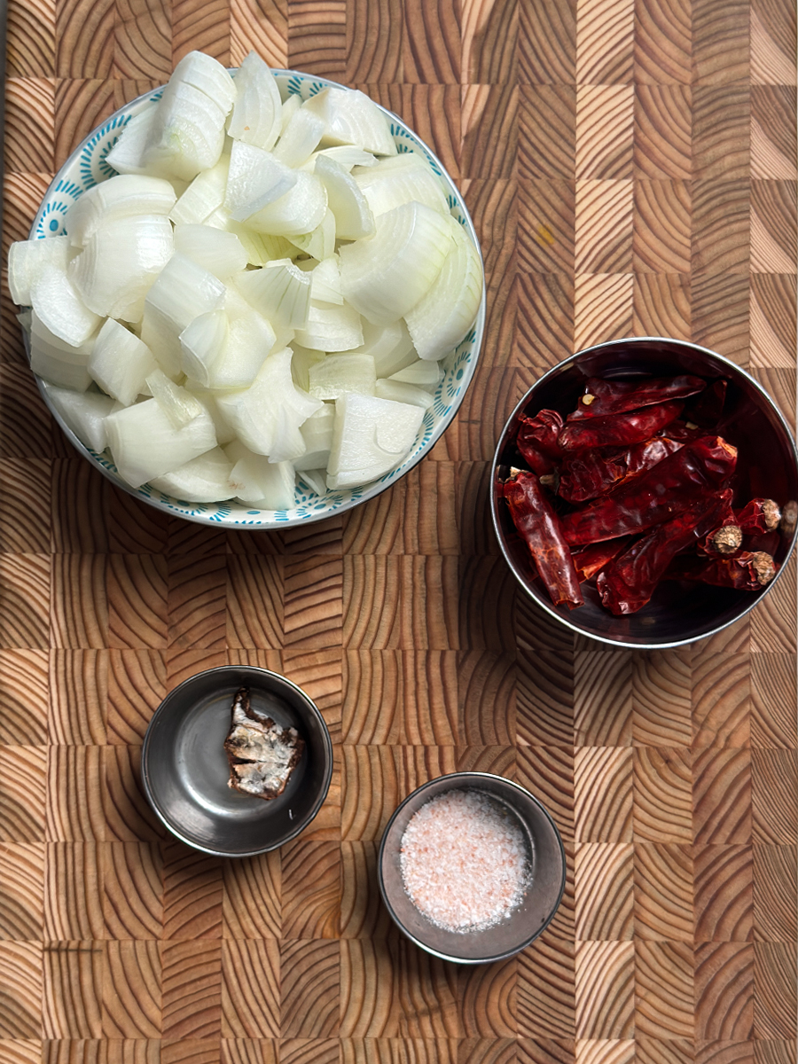 Fresh chopped white onions, dried red chilies, tamarind piece, and salt arranged in separate bowls on bamboo cutting board