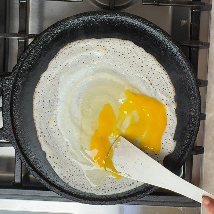 White spatula breaking bright orange egg yolk on creamy dosa surface in seasoned cast iron tawa
