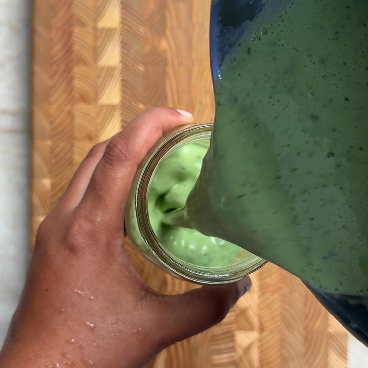 Hand holding glass jar filled with vibrant green cilantro lime yogurt dressing on wooden background