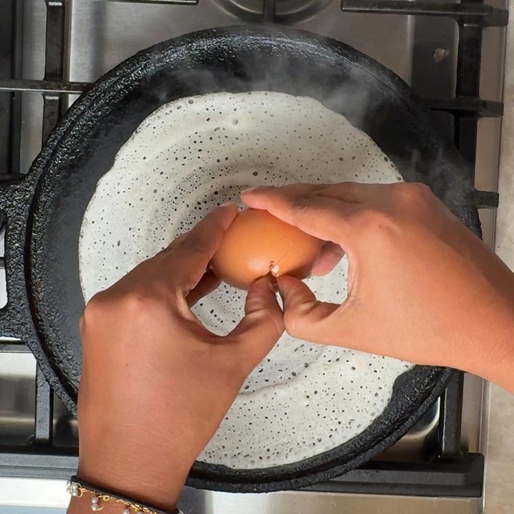 Hands cracking a brown egg over a thin white dosa with tiny bubbles cooking in black cast iron pan