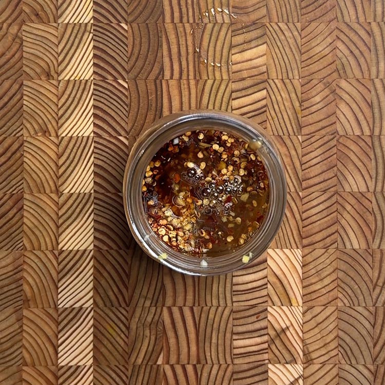 Close-up view of smooth, glossy peanut dressing showing creamy texture with scattered red pepper flakes in glass bowl