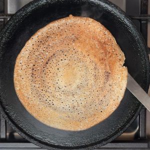 Golden brown dosa cooking in black cast iron pan with crispy texture and small air pockets