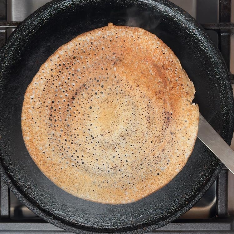 Golden brown dosa cooking in black cast iron pan with crispy texture and small air pockets