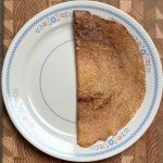 Crispy golden dosa folded in half on white plate with blue floral pattern on wooden surface