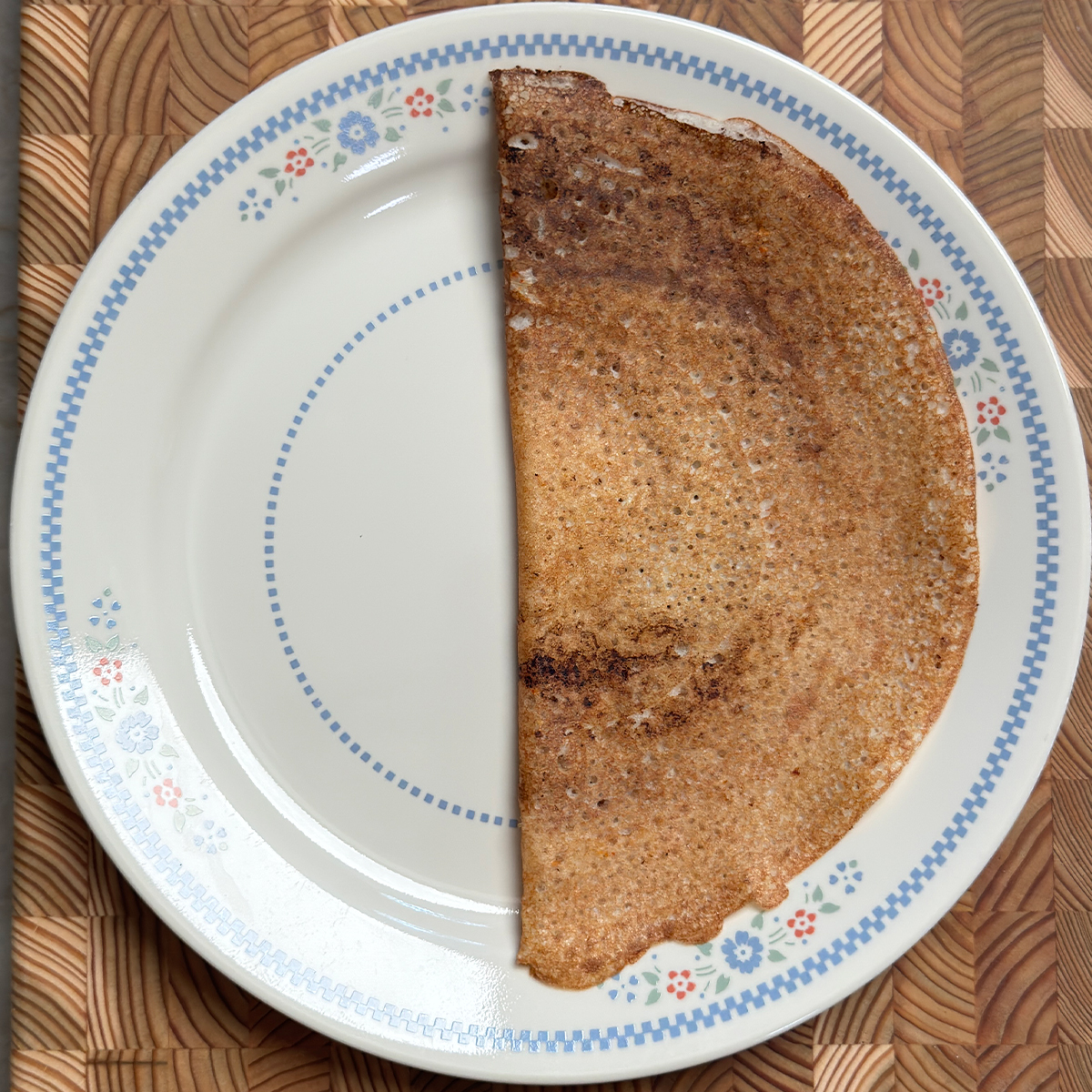 Crispy golden dosa folded in half on white plate with blue floral pattern on wooden surface