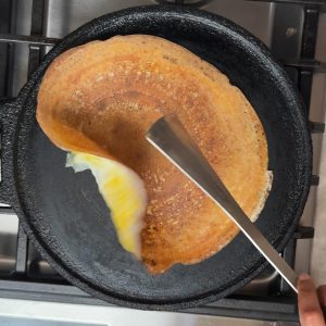Partially flipped golden dosa being turned egg-side down with spatula to cook the egg layer in cast iron pan