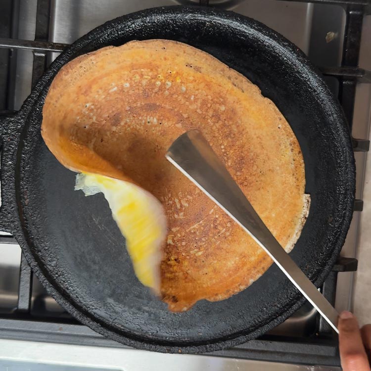 Partially flipped golden dosa being turned egg-side down with spatula to cook the egg layer in cast iron pan