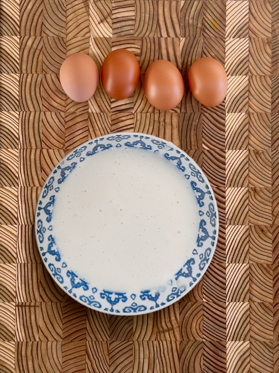Four brown eggs arranged above a blue-rimmed bowl of creamy white fermented dosa batter on bamboo mat