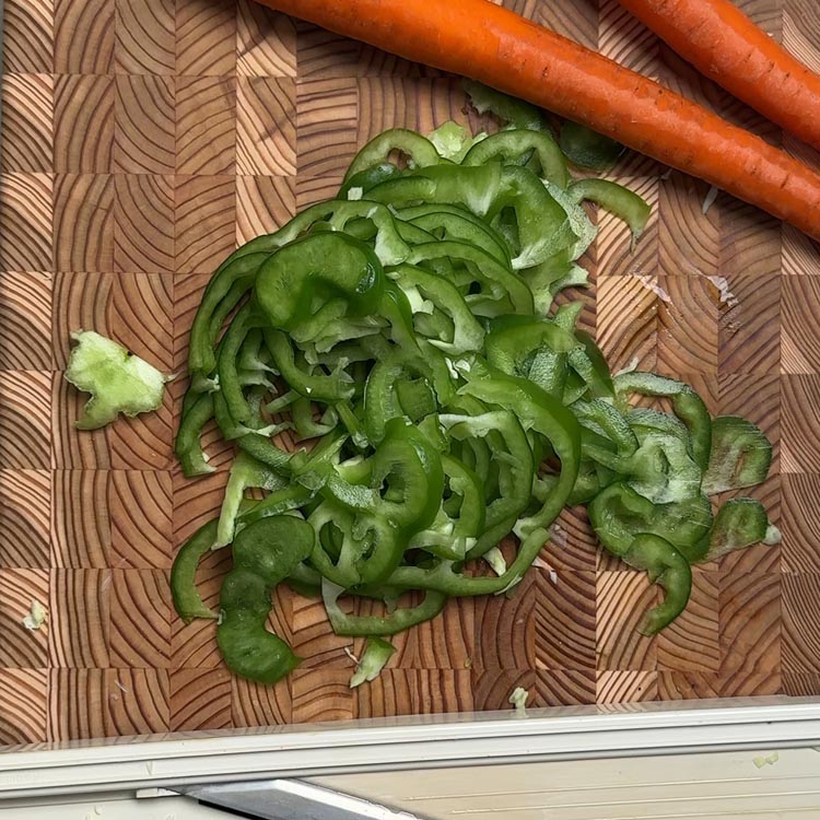Fresh green bell pepper cut into thin strips on wooden cutting board next to chef's knife