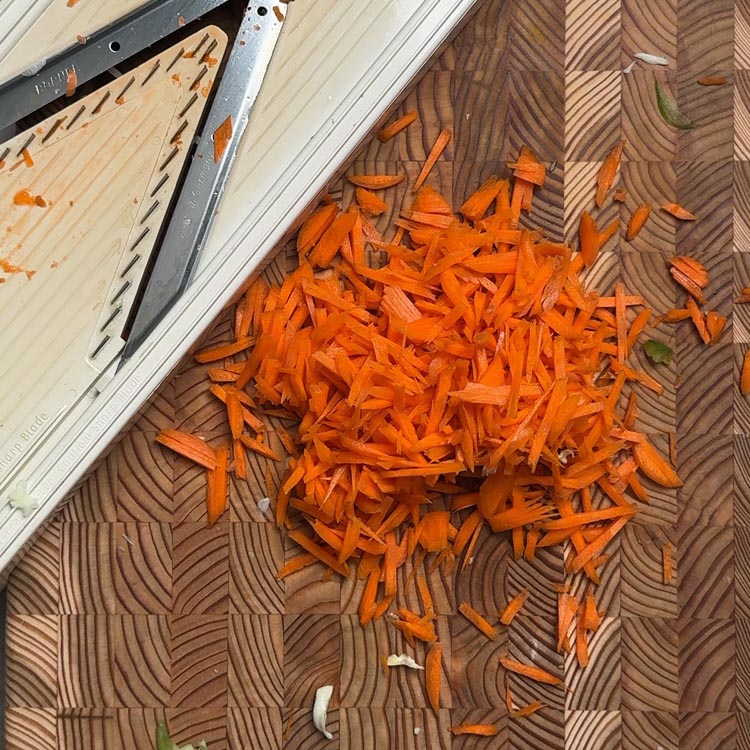 Bright orange julienned carrot strips freshly grated with mandoline slicer visible on cutting board