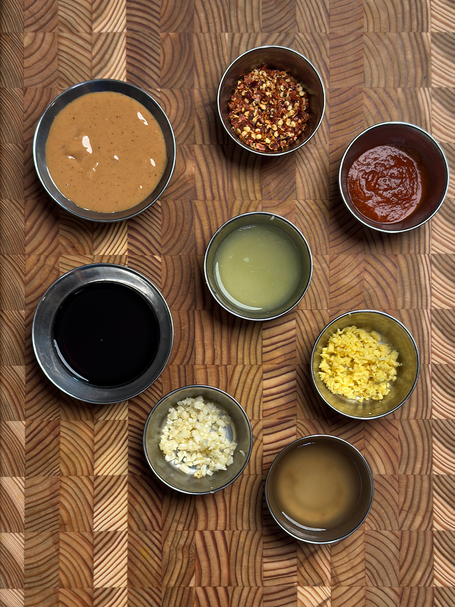Small bowls containing peanut butter, red pepper flakes, soy sauce, lime juice, rice vinegar, minced garlic and ginger arranged on textured wooden cutting board
