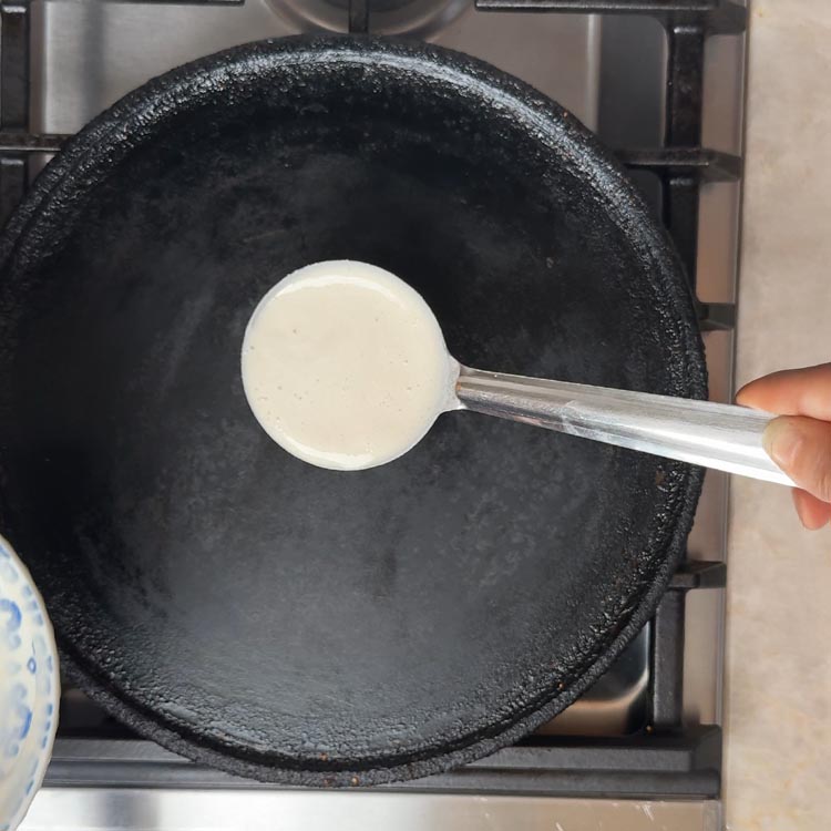 White fermented batter being poured from steel ladle onto center of black cast iron tawa