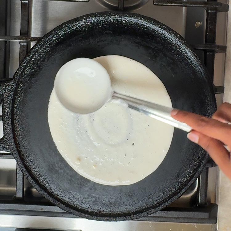 White fermented dosa batter being spread with ladle in circular motion on hot black cast iron tawa