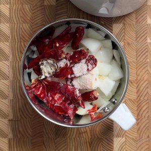 Coarse orange-red spiced paste with visible chili flecks and onion pieces in stainless steel food processor bowl