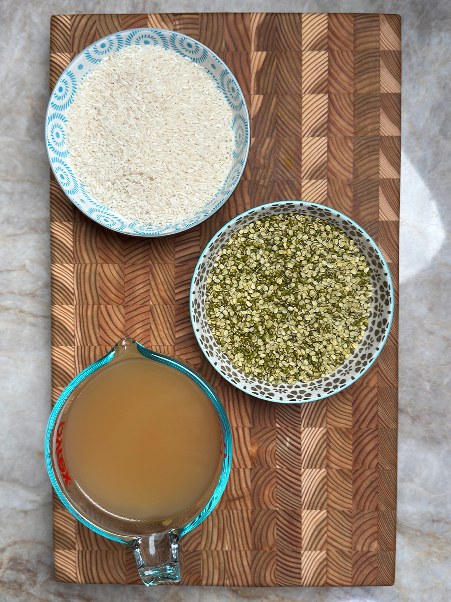 Three bowls arranged on wooden cutting board showing white rice, split green moong dal, and chicken broth