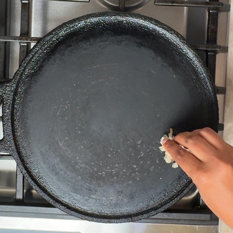 Hand rubbing cut onion piece across black cast iron tawa surface to season and prevent sticking