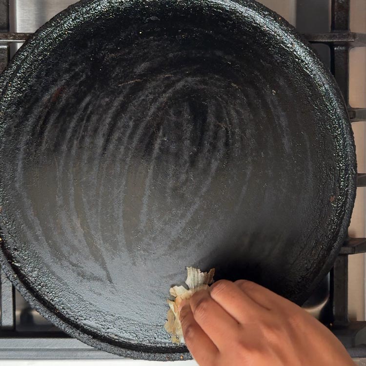 Hand rubbing a cut onion piece on hot cast iron tawa surface to prevent sticking