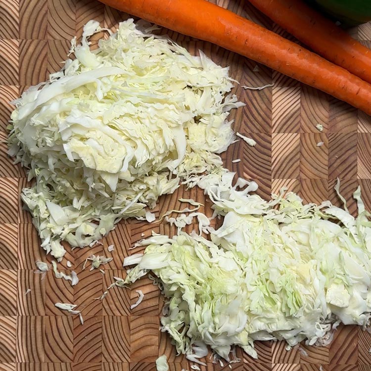 Thinly sliced white cabbage strips scattered on wooden cutting board with whole carrots in background