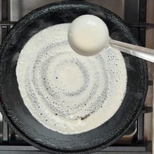 Creamy white fermented dosa batter being spread in thin circular motion on black cast iron pan