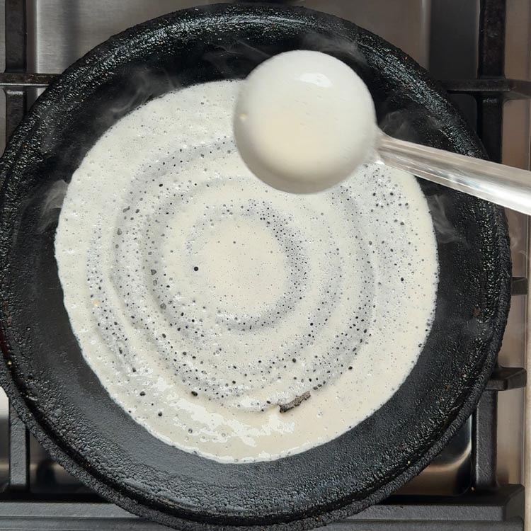 Creamy white fermented dosa batter being spread in thin circular motion on black cast iron pan