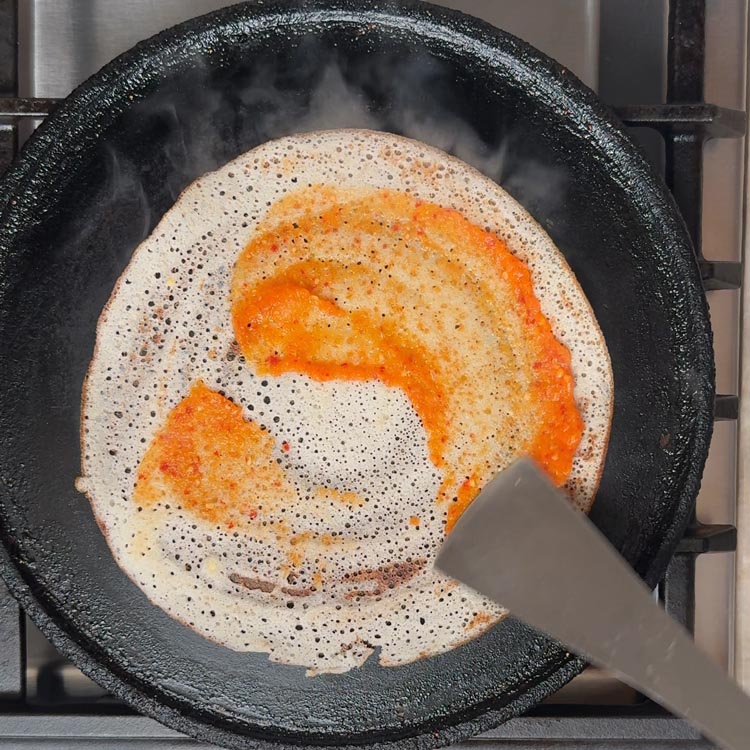 Bright red erra karam chutney being spread evenly on cooked dosa with metal spatula