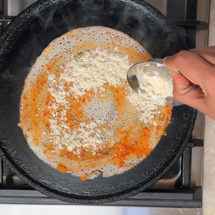 Hand sprinkling beige roasted lentil powder over red chutney spread on dosa in hot pan