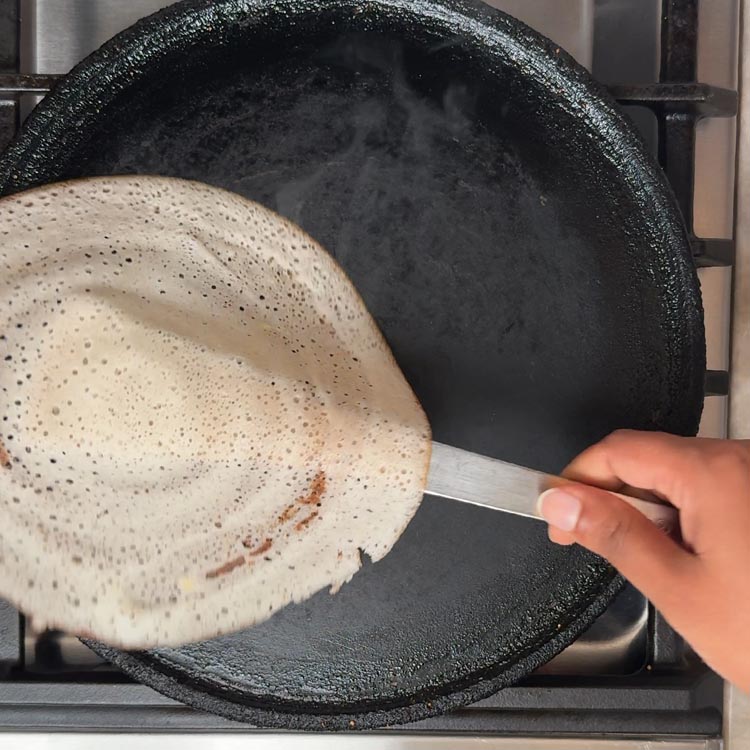 Thin white dosa crepe cooking on hot tawa with tiny fermentation bubbles visible on surface about to be flipped