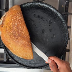 Metal spatula lifting golden brown folded dosa from cast iron pan to transfer to serving plate