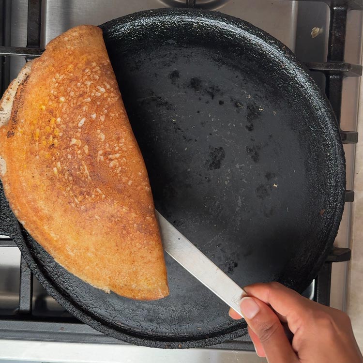 Metal spatula lifting golden brown folded dosa from cast iron pan to transfer to serving plate
