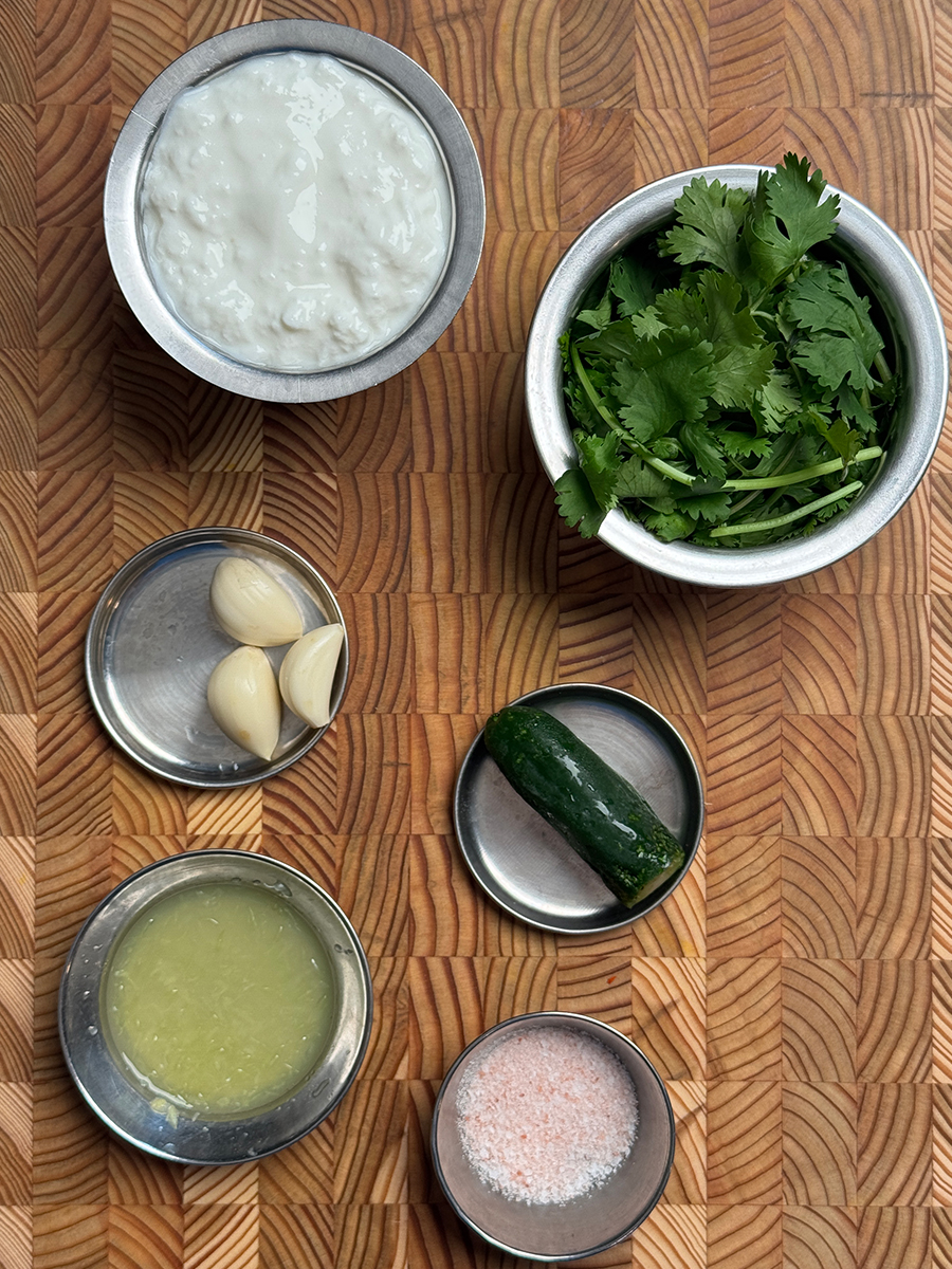 Small steel bowls containing Greek yogurt, fresh cilantro, garlic cloves, jalapeño, lime juice and salt arranged on textured wooden cutting board