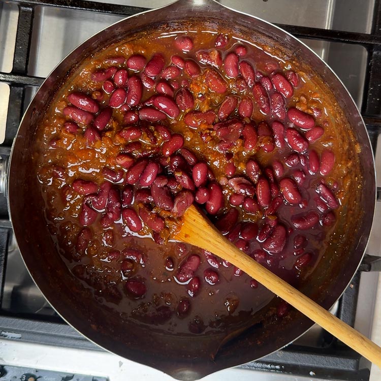 Cooked red kidney beans being stirred into bubbling brown curry base in pan