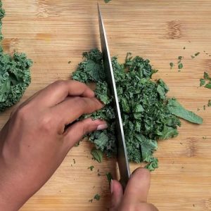 Hands using sharp knife to chop fresh green kale leaves into bite sized pieces on bamboo cutting board