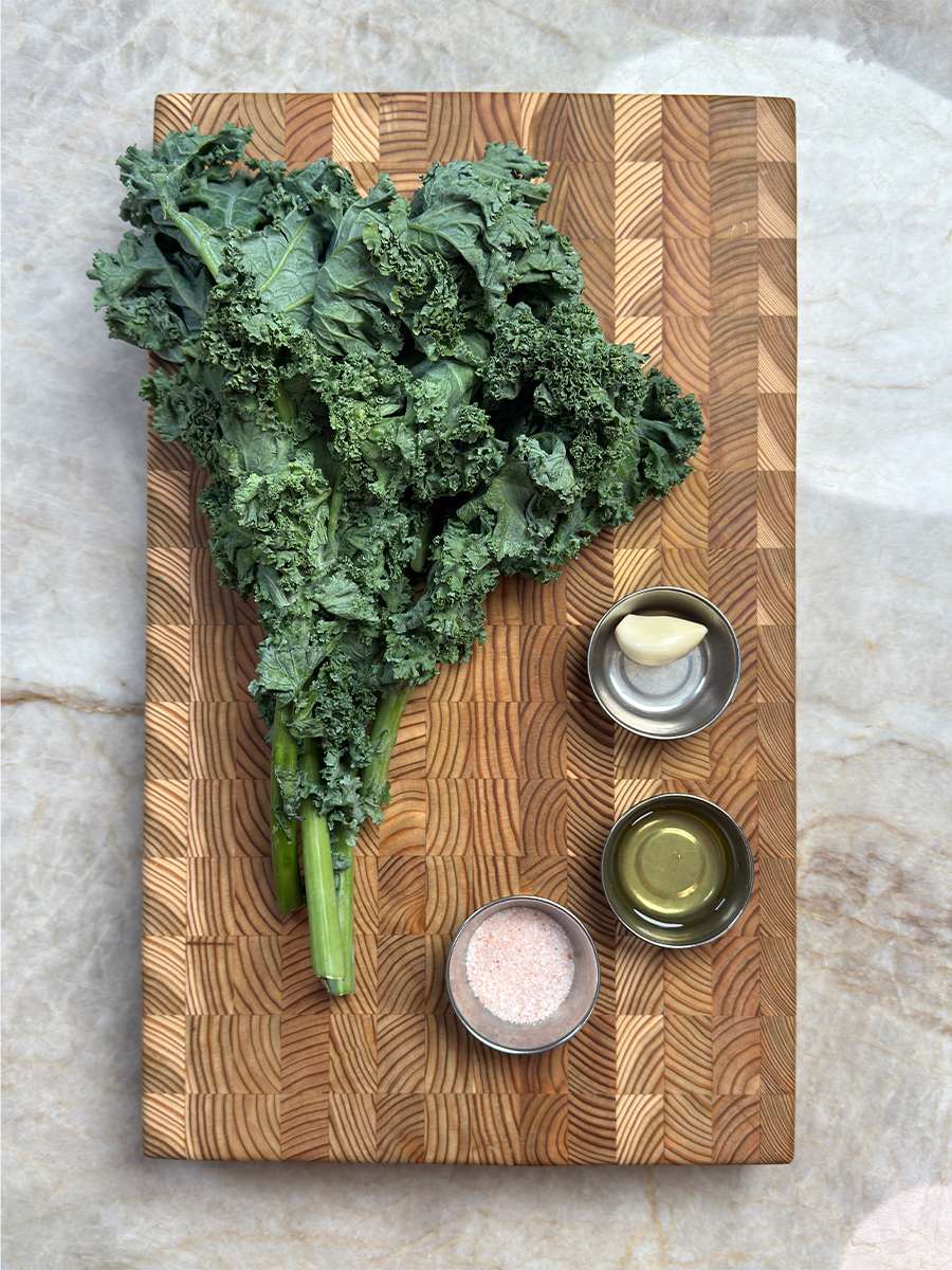 Fresh curly kale leaves with small bowls of garlic, olive oil, and salt arranged on wooden cutting board