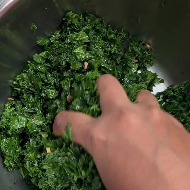 Hands massaging chopped kale with oil in large stainless steel mixing bowl, leaves becoming tender and dark