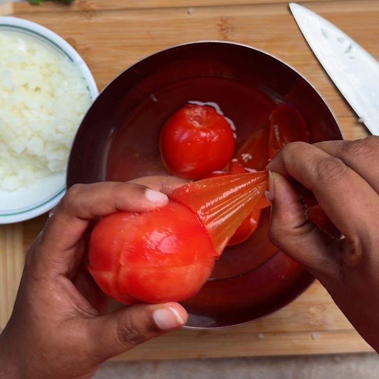 Hands peeling smooth skin from blanched red tomato in dark bowl with chopped onions nearby